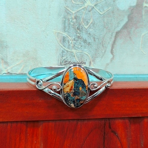 May include: A silver bangle bracelet with a large oval-shaped orange, blue, and white gemstone set in the center. The bracelet has a delicate, ornate design.