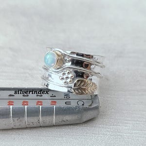 K&ouml;nnte beinhalten: Ein silberner Spinner-Ring mit einem runden, schillernden Stein, einem kleinen Blumenmotiv und einem strukturierten Blattdesign, platziert auf einem Metall-Ringma&szlig;stab, der numerische Ma&szlig;e in Millimetern anzeigt.