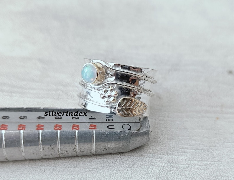 K&ouml;nnte beinhalten: Ein silberner Spinner-Ring mit einem runden, schillernden Stein, einem kleinen Blumenmotiv und einem strukturierten Blattdesign, platziert auf einem Metall-Ringma&szlig;stab, der numerische Ma&szlig;e in Millimetern anzeigt.