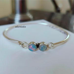 May include: A silver bangle bracelet featuring two opal stones. The bracelet has a simple design and is a delicate piece of jewellery.