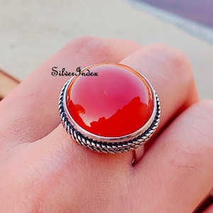 May include: A silver ring with a large, round, red carnelian gemstone. The ring has a rope-like design around the edge of the setting.