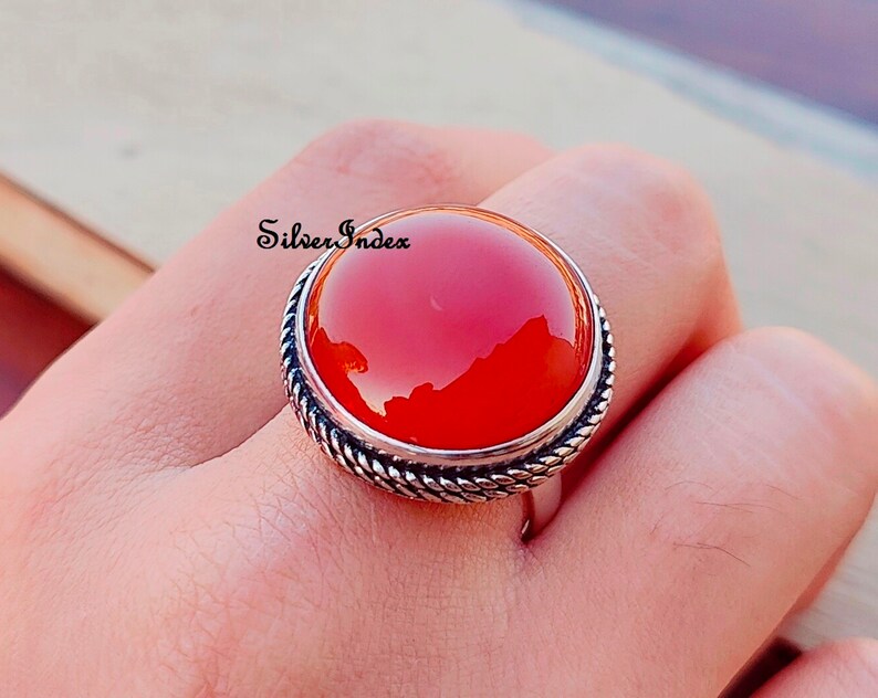May include: A silver ring with a large, round, red carnelian gemstone. The ring has a rope-like design around the edge of the setting.