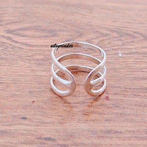 May include: A silver-colored ring with a unique open design. The ring features three bands that curve and intersect, creating an open space in the center. The word "silverindex" is printed on the ring.