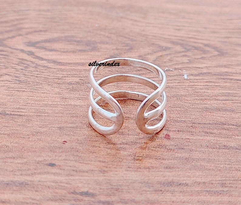 May include: A silver-colored ring with a unique open design. The ring features three bands that curve and intersect, creating an open space in the center. The word "silverindex" is printed on the ring.