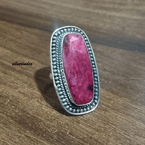 May include: A silver ring with a large, rectangular, ruby-colored stone. The stone is set in a detailed, beaded silver frame. The ring is displayed on a wooden surface.