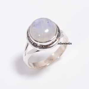 May include: A silver ring with a round, white moonstone set in a bezel setting. The ring has a simple band and is accented with a delicate silver border around the stone. "silverindex" is printed on the band.