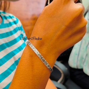 Könnte beinhalten: Ein silbernes Armreif-Armband mit floralem Muster. Das Armband befindet sich an einem Handgelenk.