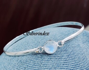 May include: A silver bangle bracelet with a round, iridescent moonstone center. The bracelet has a simple, elegant design and is displayed on a denim surface. The text "Silverindex" is visible.