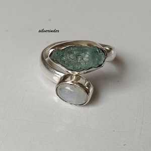 Peut inclure: Une bague en argent ornée d'une grosse pierre précieuse bleu-vert brute et d'une petite pierre précieuse ovale blanche. La bague a un design ouvert avec les pierres précieuses placées aux extrémités opposées de la bande. Le mot "silverindex" est en haut à gauche.