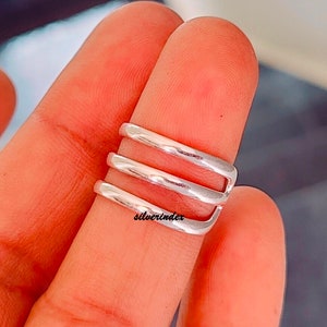 May include: A silver-colored ring with a three-band design. The ring is wrapped around a finger, showcasing its minimalist style. The ring's surface reflects light, highlighting its polished finish. The text "silverindex" is visible on the ring.