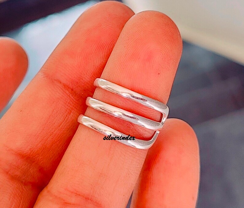 May include: A silver-colored ring with a three-band design. The ring is wrapped around a finger, showcasing its minimalist style. The ring's surface reflects light, highlighting its polished finish. The text "silverindex" is visible on the ring.