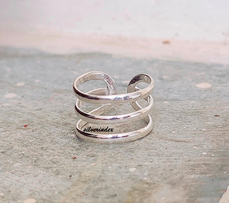 May include: A silver-colored ring with a unique design. The ring features three horizontal bands connected by a vertical loop. The word "silverindex" is engraved on the ring. The ring is made of a shiny, metallic material.