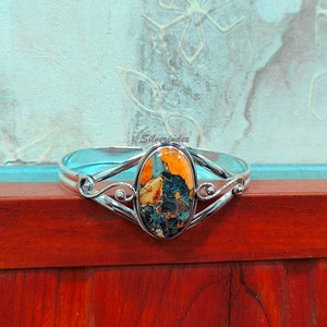 May include: A silver bangle bracelet with a large oval-shaped orange, blue, and white gemstone set in the center. The bracelet has a delicate, ornate design.