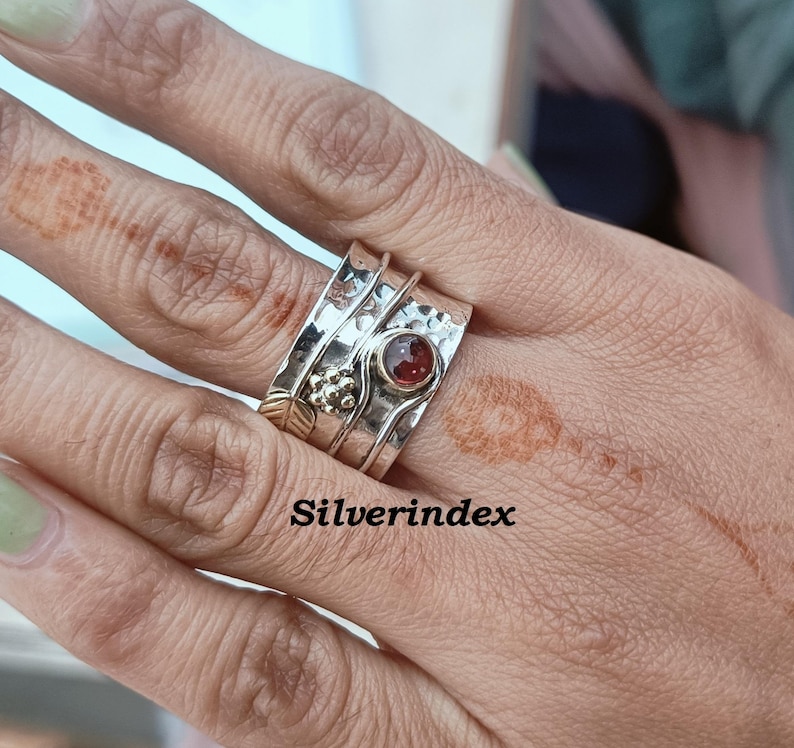 K&ouml;nnte beinhalten: Ein breiter Silberring mit Drehfunktion, geh&auml;mmertem Design, einem kleinen runden dunkelroten Edelstein, einem goldfarbenen Blattakzent und einer Ansammlung kleiner Silberperlen. Das Wort Silverindex ist in schwarzer Schrift auf dem Ring zu sehen.