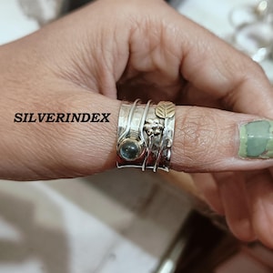 K&ouml;nnte beinhalten: Ein breiter Silberring mit Drehfunktion, geh&auml;mmertem Design, einem runden blauen Edelstein sowie goldfarbenen Akzenten in Form einer Blume und eines Blattes. Der Schriftzug SILVERINDEX ist links auf dem Ringband zu sehen, das am Daumen getragen wird.