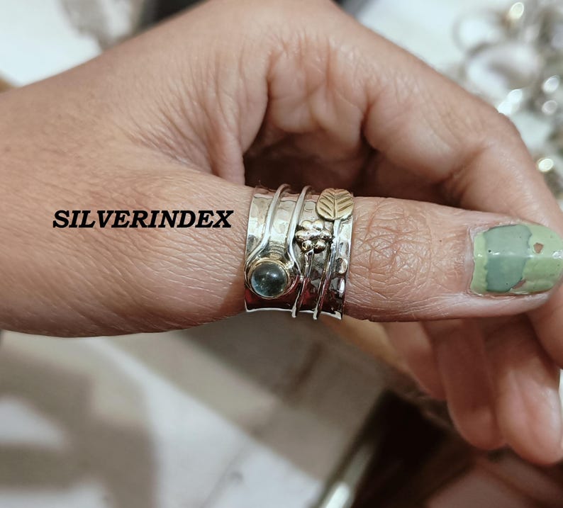 K&ouml;nnte beinhalten: Ein breiter Silberring mit Drehfunktion, geh&auml;mmertem Design, einem runden blauen Edelstein sowie goldfarbenen Akzenten in Form einer Blume und eines Blattes. Der Schriftzug SILVERINDEX ist links auf dem Ringband zu sehen, das am Daumen getragen wird.