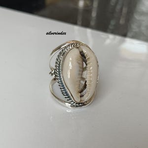 Puede incluir: Un anillo de plata con un centro de concha de cauri natural. La concha es de color blanco cremoso, engastada en una banda de plata detallada con un diseño texturizado. El anillo tiene una estética única y orgánica.
