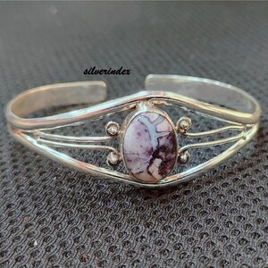 May include: A silver bangle bracelet with a purple and white stone set in the center. The bracelet has a delicate, intricate design.