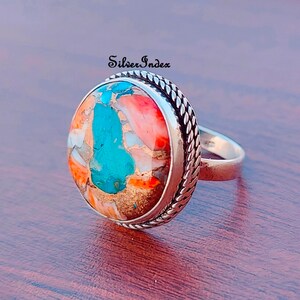 May include: A silver ring with a large, oval-shaped turquoise and coral stone set in a rope-like border.