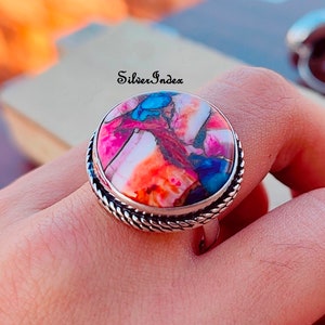 May include: A silver ring with a large, round, multi-colored stone set in a silver bezel. The stone has shades of pink, orange, blue, and brown. The ring has a rope-like design around the band.