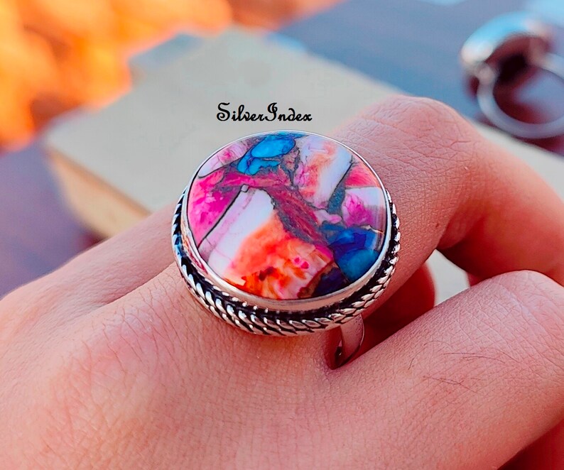 May include: A silver ring with a large, round, multi-colored stone set in a silver bezel. The stone has shades of pink, orange, blue, and brown. The ring has a rope-like design around the band.