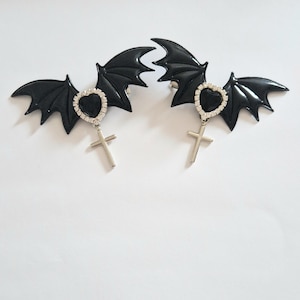 Puede incluir: Dos pinzas para el pelo con forma de alas de murciélago negras. Cada pinza presenta un colgante de corazón con pedrería y un dije de cruz plateada. Las alas son brillantes y detalladas, con una estética gótica o alternativa. Fondo blanco.
