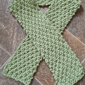 Może przedstawiać: Zielony szalik wykonany na szydełku z fakturowanym wzorem. Szalik jest złożony na pół i leży płasko na powierzchni wyłożonej płytkami.