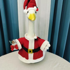 May include: A plush white goose wearing a Santa Claus outfit. The outfit includes a red jacket with white trim, a black belt with a gold buckle, and a matching Santa hat. The goose is holding a small gift.