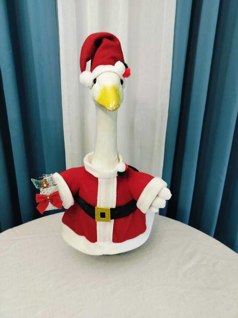 May include: A plush white goose wearing a Santa Claus outfit. The outfit includes a red jacket with white trim, a black belt with a gold buckle, and a matching Santa hat. The goose is holding a small gift.