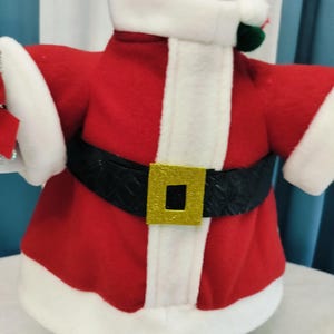 May include: A festive Santa Claus costume for a decorative item. The outfit features a red jacket with white trim, a black belt with a gold buckle, and white cuffs. The costume is designed to fit around a cylindrical object.