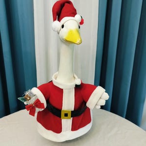May include: A plush goose wearing a Santa Claus outfit. The goose has a white body, a yellow beak, and is dressed in a red jacket with white trim, a black belt with a gold buckle, and a red Santa hat. It is holding a small decorative item.