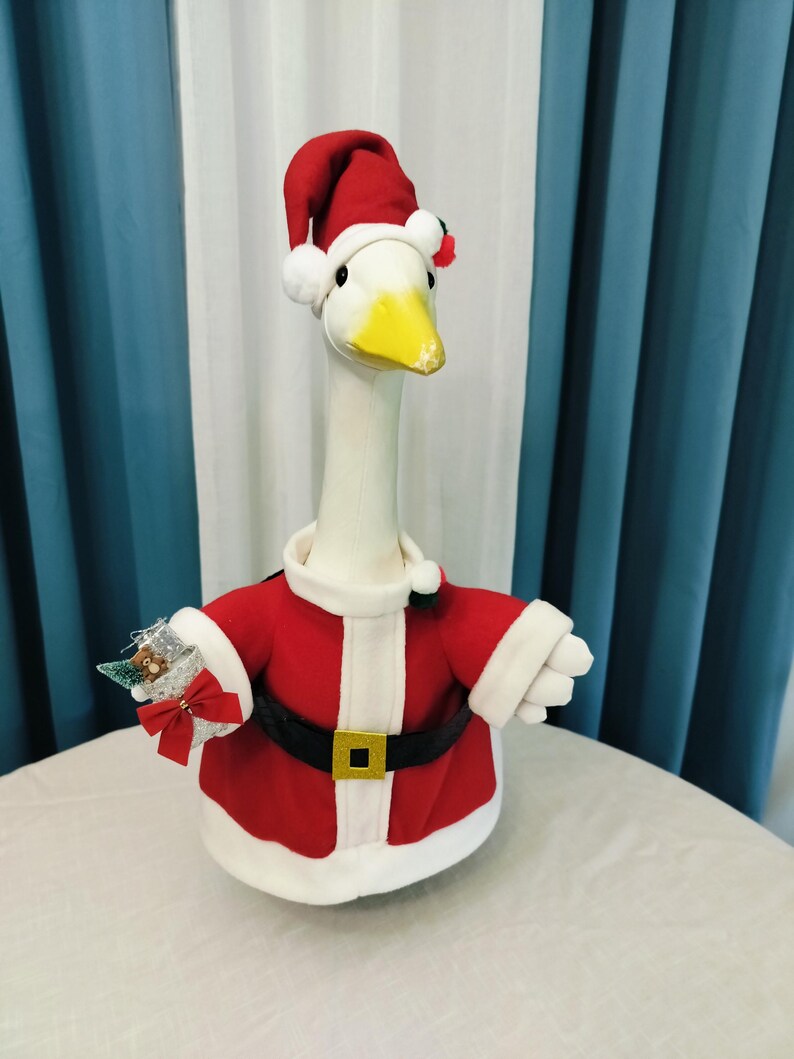 May include: A plush goose wearing a Santa Claus outfit. The goose has a white body, a yellow beak, and is dressed in a red jacket with white trim, a black belt with a gold buckle, and a red Santa hat. It is holding a small decorative item.