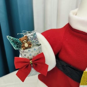 May include: A festive Santa Claus figure with a red suit, white trim, and a black belt. The figure holds a silver glittery container adorned with a red bow, a small teddy bear, a miniature Christmas tree, and a wrapped gift.