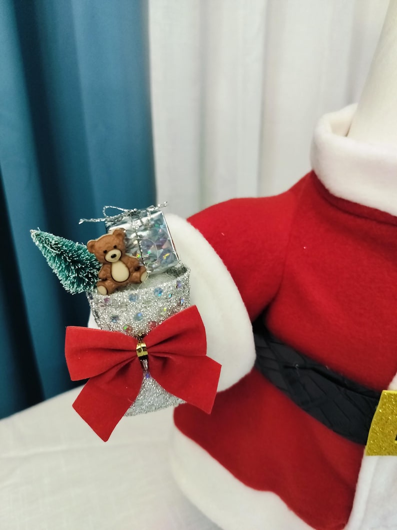 May include: A festive Santa Claus figure with a red suit, white trim, and a black belt. The figure holds a silver glittery container adorned with a red bow, a small teddy bear, a miniature Christmas tree, and a wrapped gift.