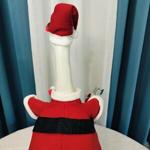 May include: A white goose figurine dressed in a red Santa Claus outfit, complete with a matching hat and black belt. The goose is wearing a red coat with white trim and holding a small decorative item. The background features white and blue curtains.