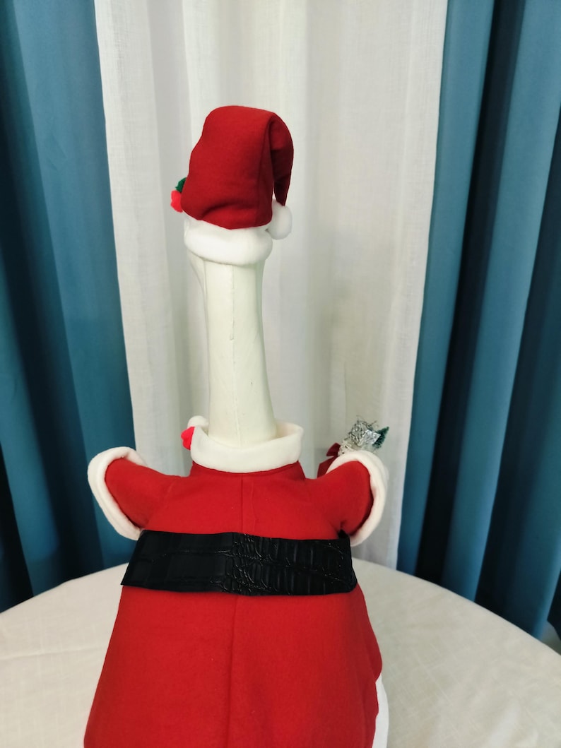May include: A white goose figurine dressed in a red Santa Claus outfit, complete with a matching hat and black belt. The goose is wearing a red coat with white trim and holding a small decorative item. The background features white and blue curtains.