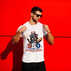 Op de afbeelding: Een wit t-shirt met een afbeelding van een wasbeer die een patriottische hoge hoed, een zonnebril met sterren en een bandana draagt. De wasbeer houdt een sterretje en een hotdog vast. Onderaan staat 250 YEARS OF QUESTIONABLE DECISIONS.