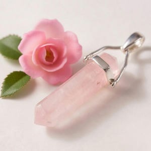 May include: A pink rose quartz crystal pendant with a silver-colored bail. The pendant is next to a pink flower with green leaves. The crystal is a light pink color and has a pointed end.