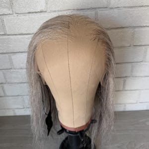 Könnte beinhalten: Eine lange, gerade, silbergraue Perücke auf einem Mannequin-Kopf. Die Perücke ist mit einem Mittelscheitel gestylt und hat eine natürliche, leicht gewellte Textur.