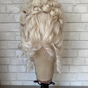 Könnte beinhalten: Hellblonde Perücke mit großen, gestylten Locken, verziert mit einem floralen und perlenbesetzten Haarschmuck. Die Perücke wird auf einem Schaufensterpuppenkopf präsentiert. Der Hintergrund ist eine weiße Backsteinmauer. Die Locken umrahmen das Gesicht.