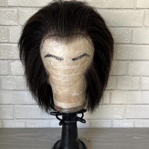 Könnte beinhalten: Eine schwarze Perücke mit einem Spitzenfront auf einem Perückenständer. Die Perücke ist in einem kurzen, geraden Bob gestylt.