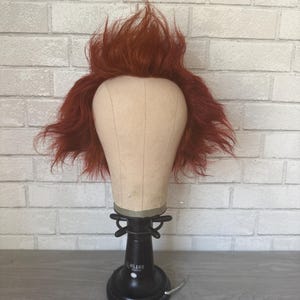 May include: A red wig with a messy, spiky style sits on a beige wig stand. The stand is mounted on a black base with the word "MILANO" printed on it. The background is a white brick wall.