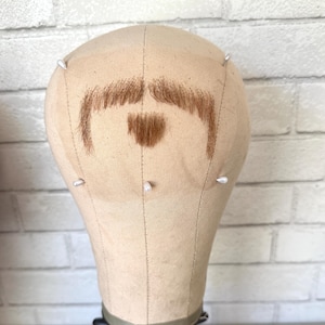 May include: A beige wig head with a brown mustache and goatee made of hair. The wig head is secured with white pins and sits on a black stand. The background is a white brick wall.
