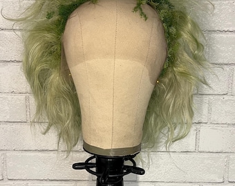 May include: A light green wig with long, wavy hair is displayed on a mannequin head. The wig is adorned with a crown of green moss. The mannequin head is mounted on a black stand, set against a white brick wall.