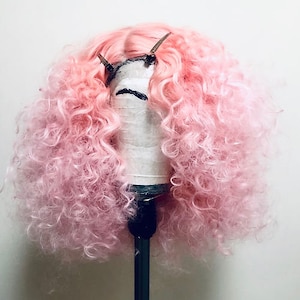 May include: A pink curly wig on a wig stand. The wig is styled in a voluminous, curly fashion.
