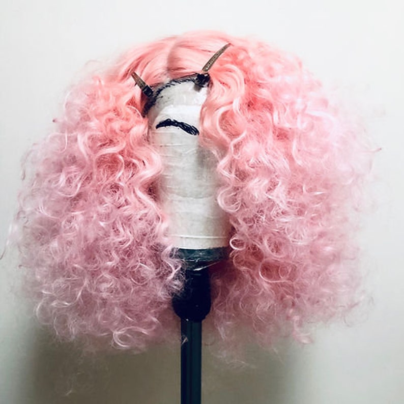 Puede incluir: Una peluca rizada rosa en un soporte para pelucas. La peluca est&aacute; peinada con un estilo voluminoso y rizado.