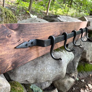 May include: A rectangular wooden board with a dark brown stain featuring a horizontal black metal bar attached to the front. The bar has a pointed arrow-shaped end and four curved metal hooks extending downward for hanging items.
