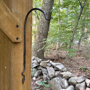 May include: A black metal plant hook with a curved top and a decorative leaf-shaped finial at the bottom, mounted to a vertical wooden post with two visible screws. The hook features a slender, hand-forged appearance with a dark, textured finish.