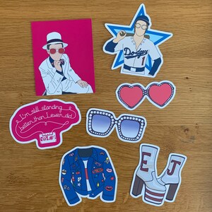 May include: A set of 10 colourful stickers featuring a baseball player, sunglasses, a jacket, a cassette tape, a heart, and a star. The stickers are printed on white paper with black outlines.