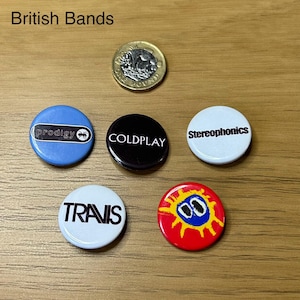 May include: Five round, colourful buttons with the names of British bands: Prodigy, Coldplay, Stereophonics, Travis, and a red button with a yellow sun and blue eyes.